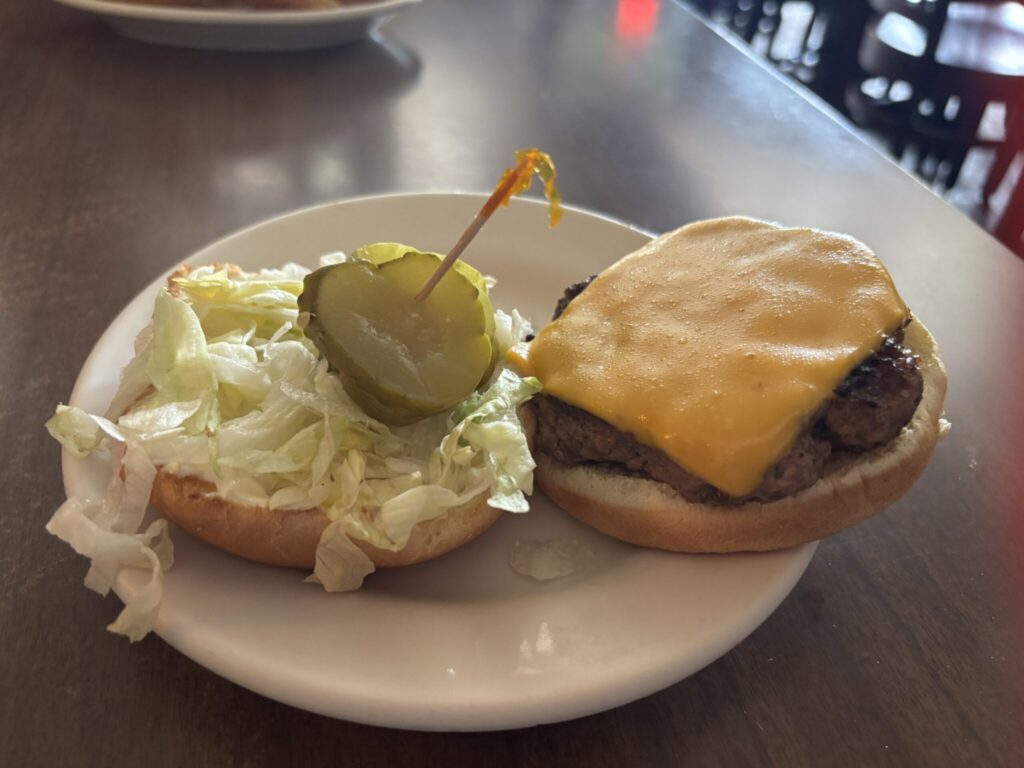 A burger from Zip's Cafe. THe burger is topped with american cheese and lettuce and pickle with a yellow topped tooth pick in the top.