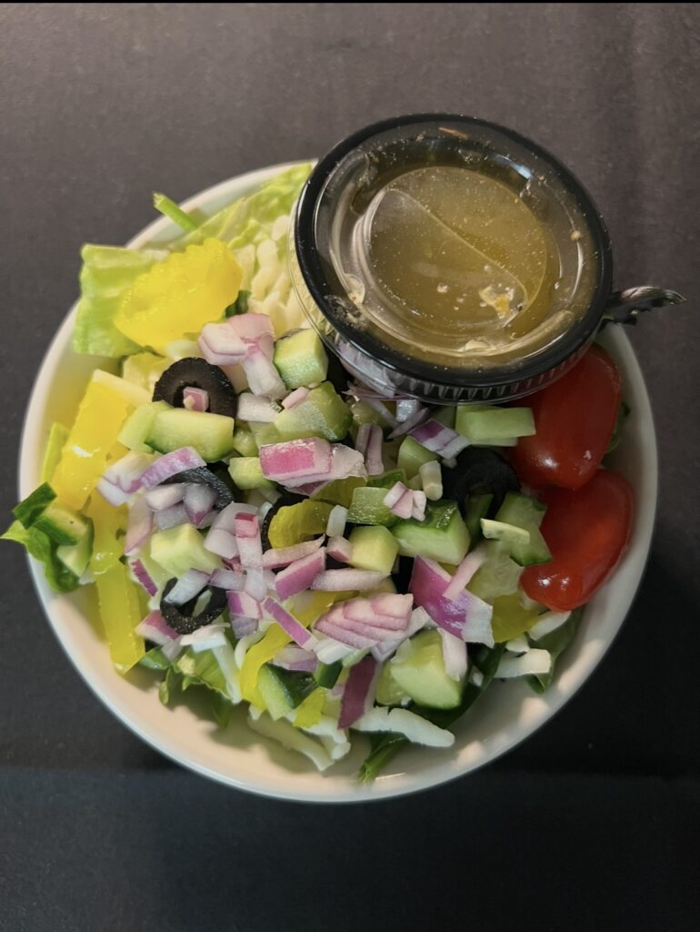 A salad of mixed greens topped with diced cucumber, tomato, red onion, black olive, and banana peppers with a side of Italian dressing.