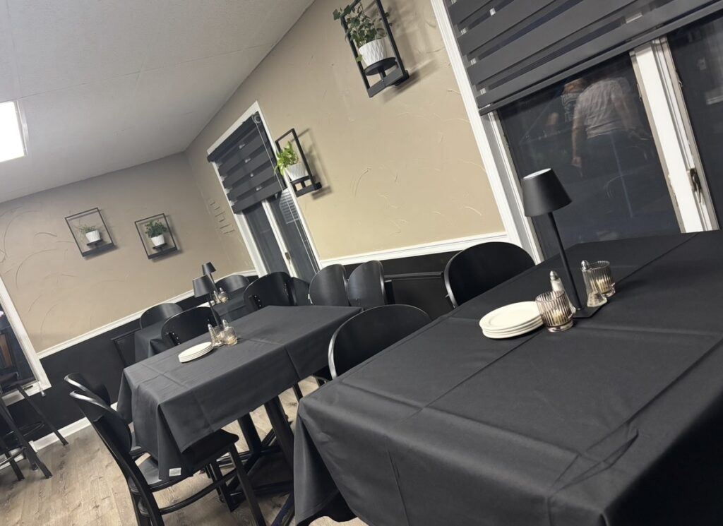 Tables in a restaurant with black tablecloths and black chairs with plants in rod iron holders hanging on the tan walls. Windows also having black blinds.