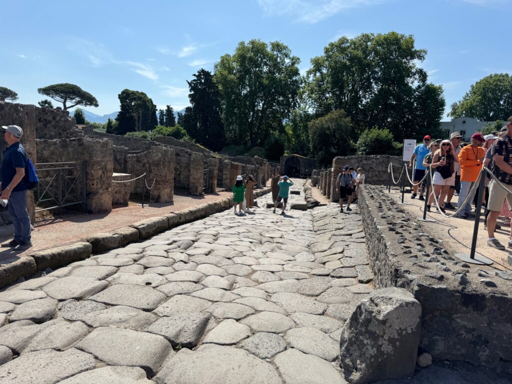 Tourists walk through ancient Roman ruins with stone buildings and a paved street under sunny skies