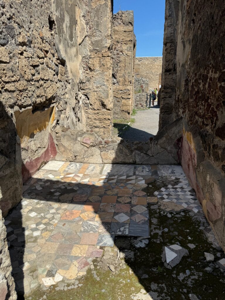 Narrow ancient stone alleyway with patterned floor tiles and weathered walls, tourists visible in distance under clear sky