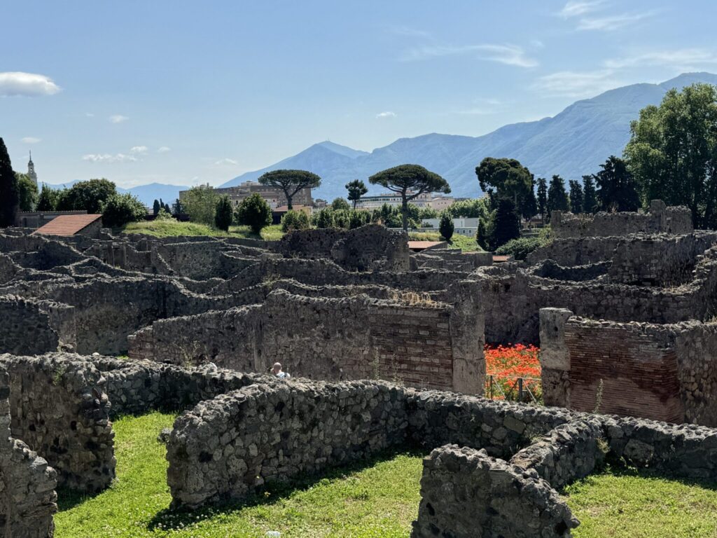 Ancient Roman ruins with stone walls in foreground, lush green landscape, and Mount Vesuvius visible in distant background