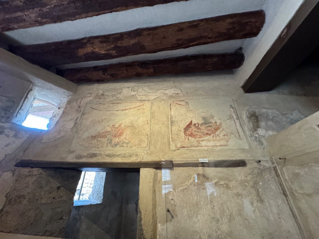 Ancient Roman frescoes with faded red ochre figures on cream-colored walls beneath exposed wooden ceiling beams in archaeological site