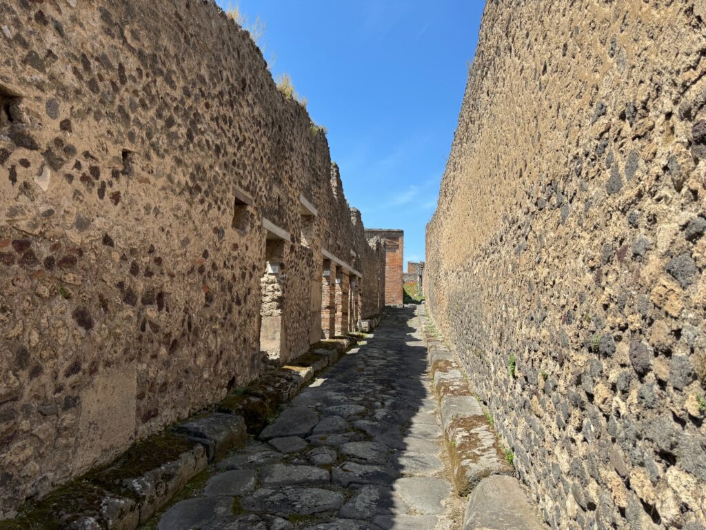 Narrow stone-paved alleyway lined with ancient volcanic rock walls under clear blue sky, likely from Pompeii or similar Roman archaeological site