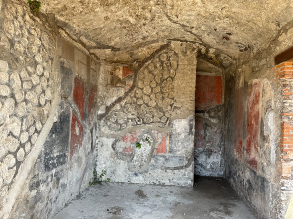 Deteriorating ancient Roman interior room with faded red walls, geometric mosaics, and crumbling plaster surfaces