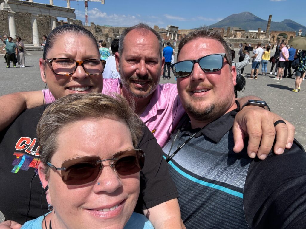 Four people smile at the camera at Pompeii archaeological site with ancient ruins and Mount Vesuvius visible in background