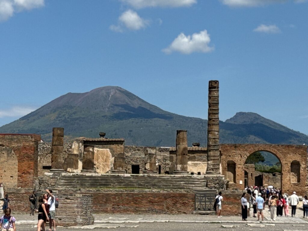 Ancient Roman ruins with standing columns and brick structures in Pompeii, with Mount Vesuvius looming in the background under a blue sky