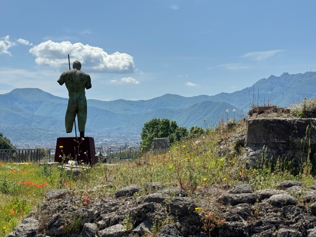 Bronze statue of a nude male figure stands on a pedestal overlooking a mountainous valley landscape