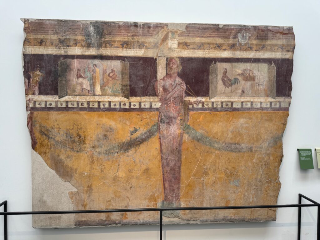 Ancient Roman fresco fragment displaying architectural scenes, figures, and decorative elements on weathered plaster in museum display