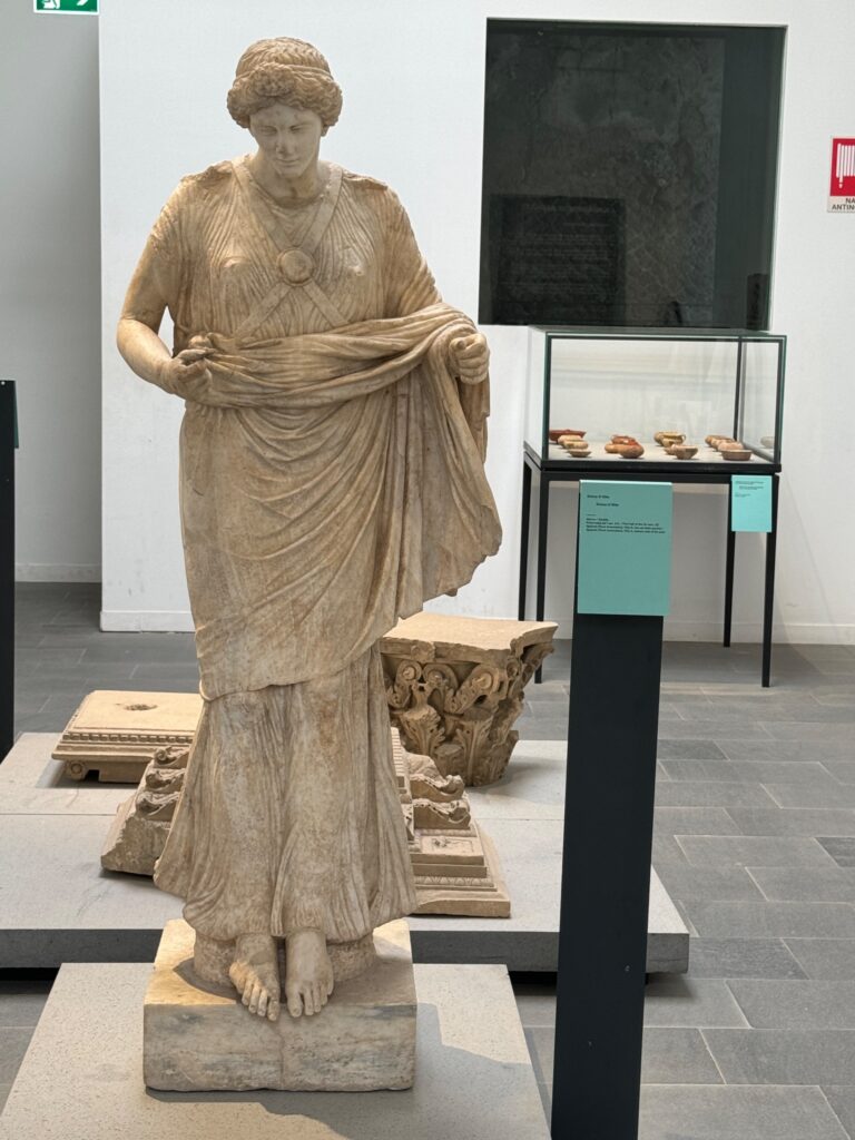 Classical marble statue of a robed female figure in museum gallery with decorative architectural fragments and display case