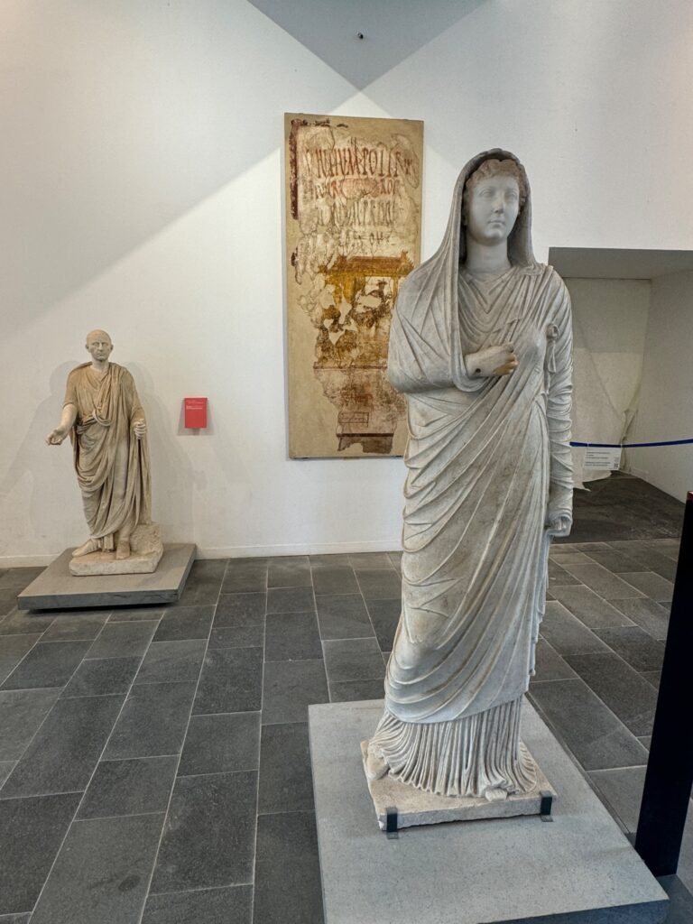 Museum gallery displaying classical Roman statues including a draped female figure and a robed male figure, with an ancient inscribed tablet on the wall