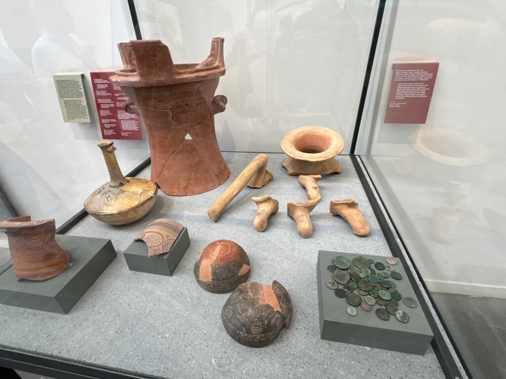 Museum display case containing ancient pottery vessels, tools, coins, and artifacts arranged on a gray platform with informational plaques