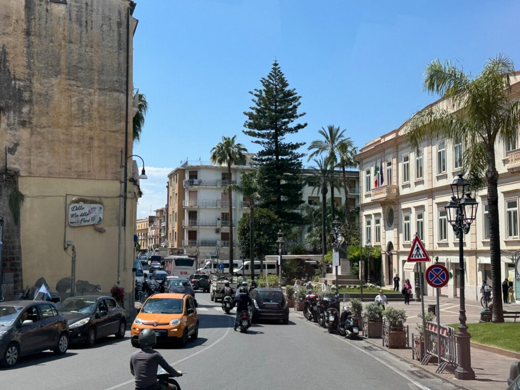 A sunny Mediterranean street scene with historic buildings, palm trees, a yellow taxi, and pedestrians in an Italian city