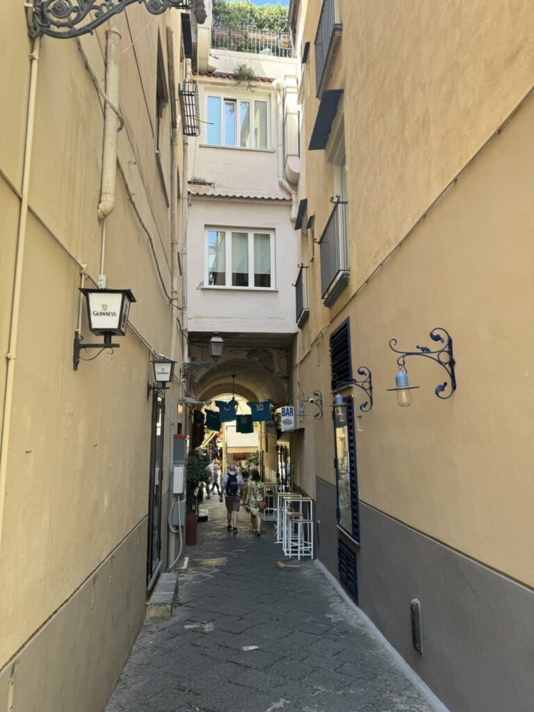 Narrow European alleyway with tall yellow buildings, shops, and pedestrians walking through an arched passage