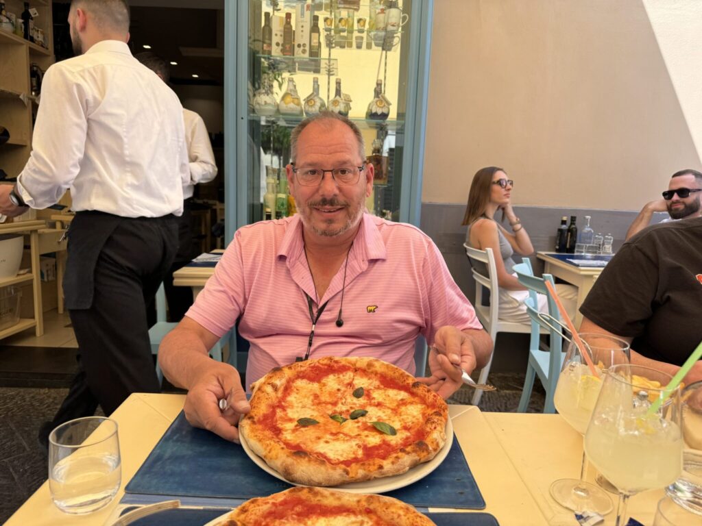 A man in a pink shirt and glasses sits at a restaurant table with a pizza in front of him, smiling at the camera