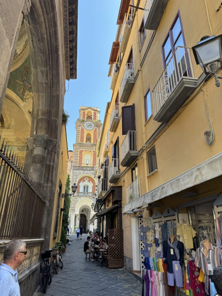Narrow European street with yellow buildings, shops, and a colorful bell tower visible in the distance under clear blue sky