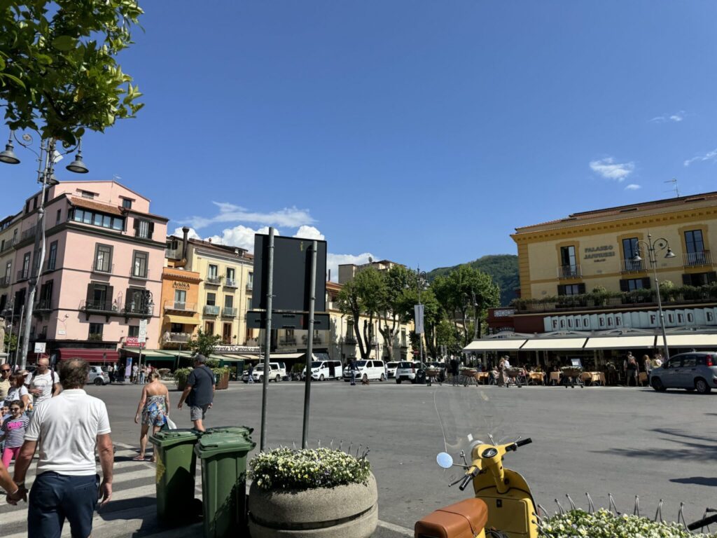 A vibrant European plaza with colorful historic buildings, pedestrians, outdoor cafes, and a yellow scooter in the foreground under a clear blue sky