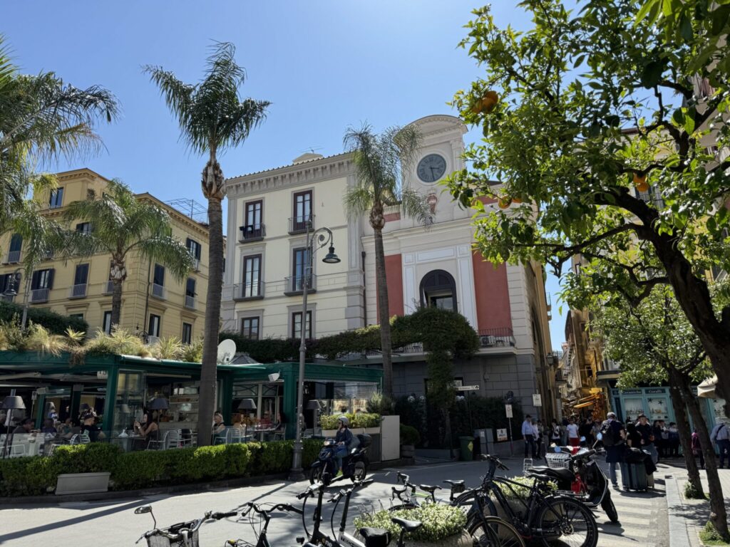 A sunny Mediterranean plaza with historic buildings, palm trees, bicycles, and pedestrians gathered in front of a cream and red-trimmed building with a clock tower