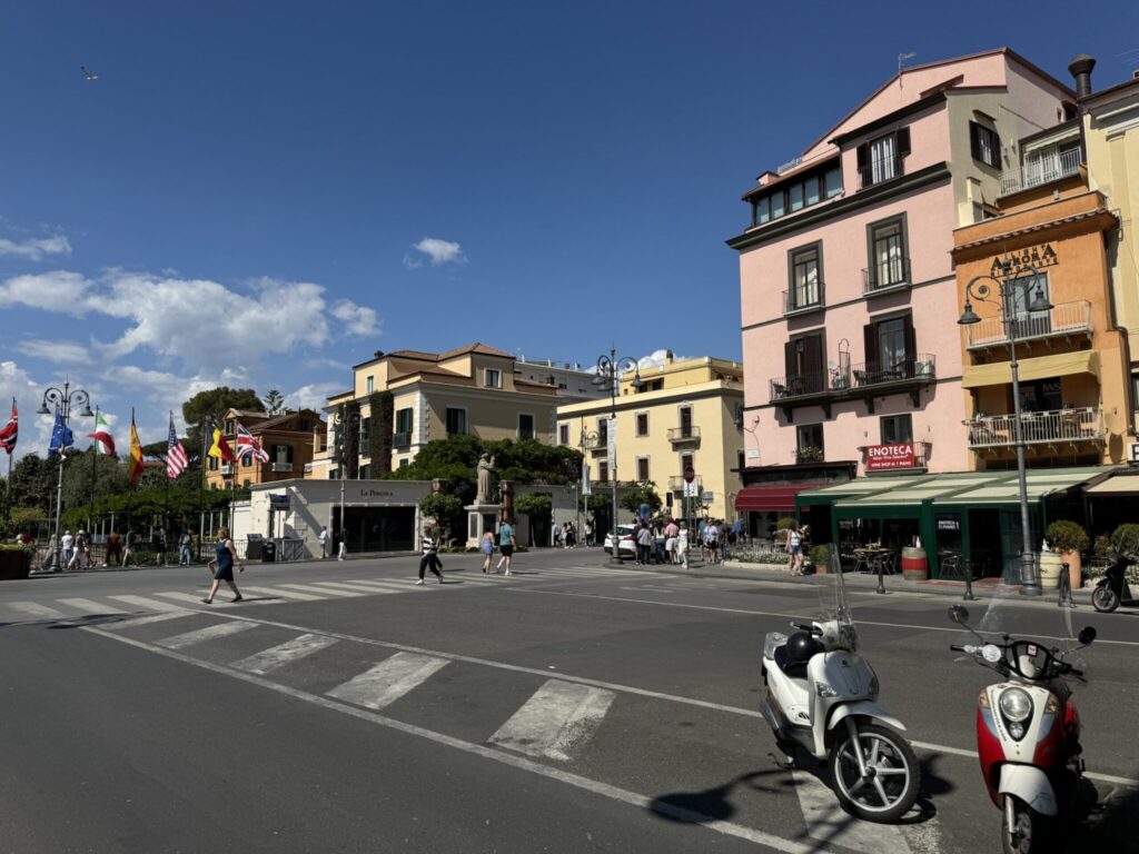 Colorful historic buildings frame a sunny Italian piazza with scooters, pedestrians, and cafes