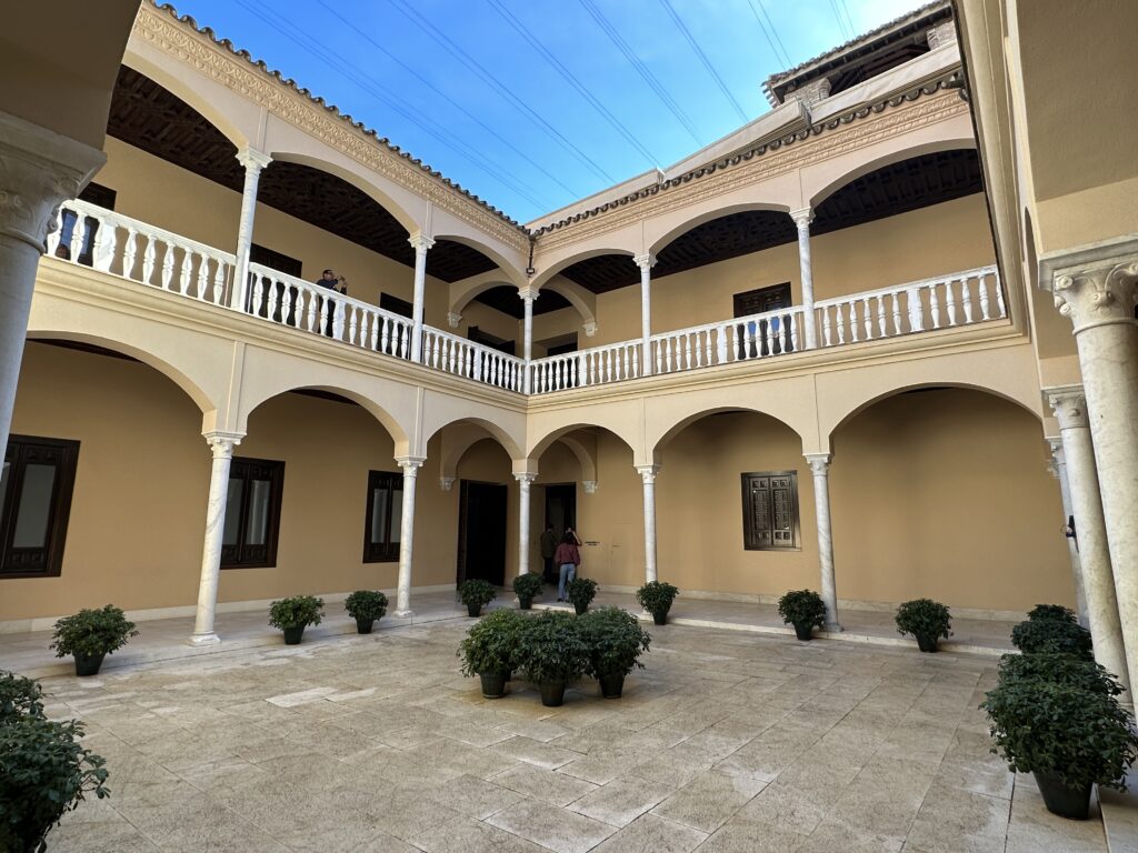 A symmetrical courtyard with cream-colored walls, arched colonnades, white railings, and potted plants under clear blue sky