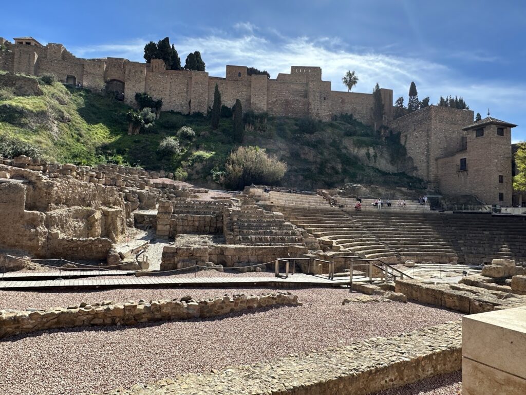 Ancient Roman theater ruins with Alcázar fortress on hillside in Málaga, Spain, under blue sky
