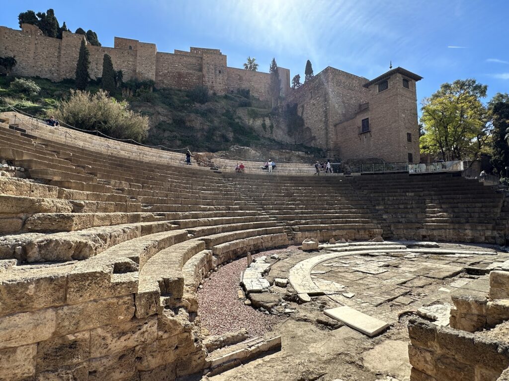 Roman amphitheater ruins with stone seating and fortified castle walls on hillside under blue sky