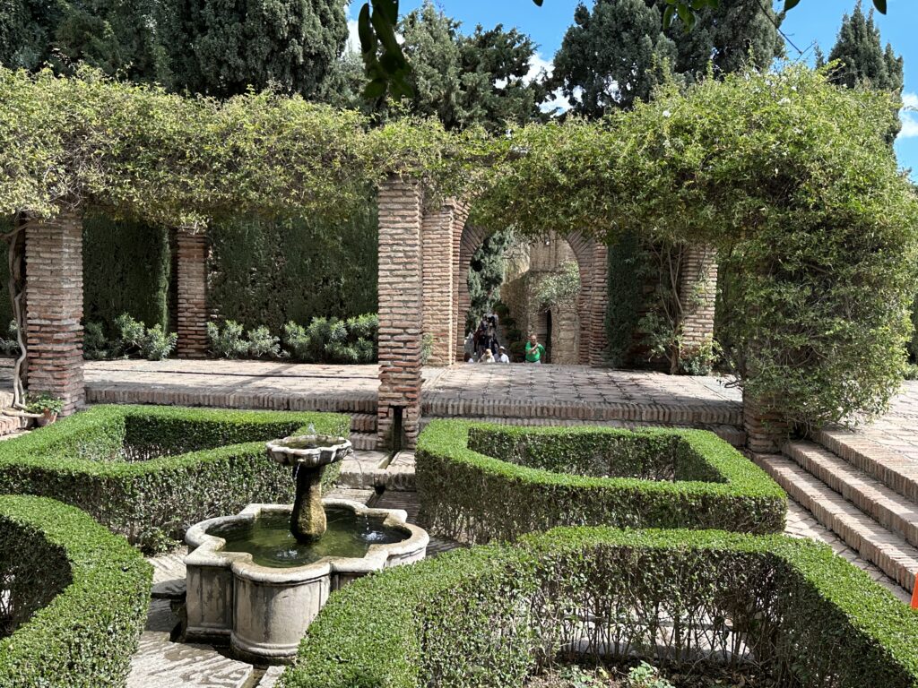 A formal garden features manicured hedges, a central fountain, and ivy-covered brick archways under tall Mediterranean trees