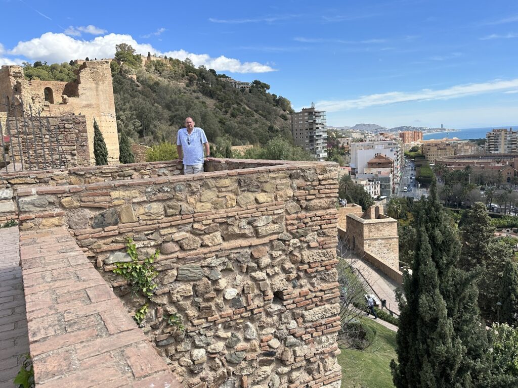 Man standing on ancient stone fortress walls overlooking Mediterranean coastal city with modern buildings and sea in background