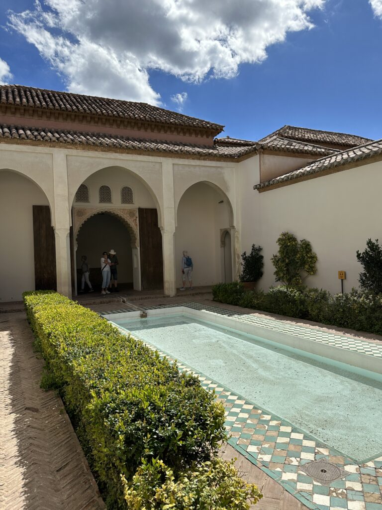 A traditional Moroccan riad courtyard features white arched walls, tiled fountain, manicured hedges, and ornate clay tile roofing