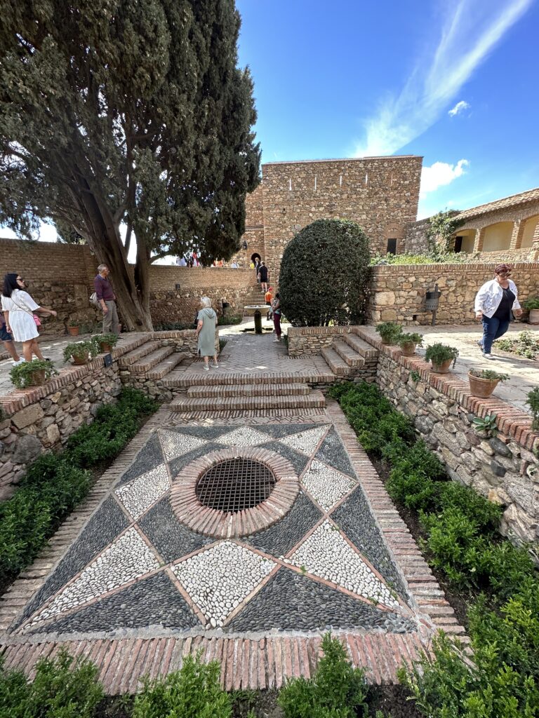 A courtyard in a historic stone fortress features geometric patterned flooring, a tall cypress tree, and medieval architecture under clear blue sky
