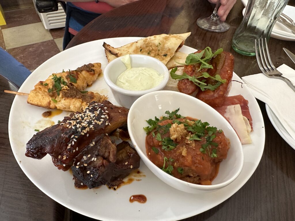 A white plate of grilled meats including skewered chicken and glazed beef with sesame seeds, served with dips and bread at a restaurant table