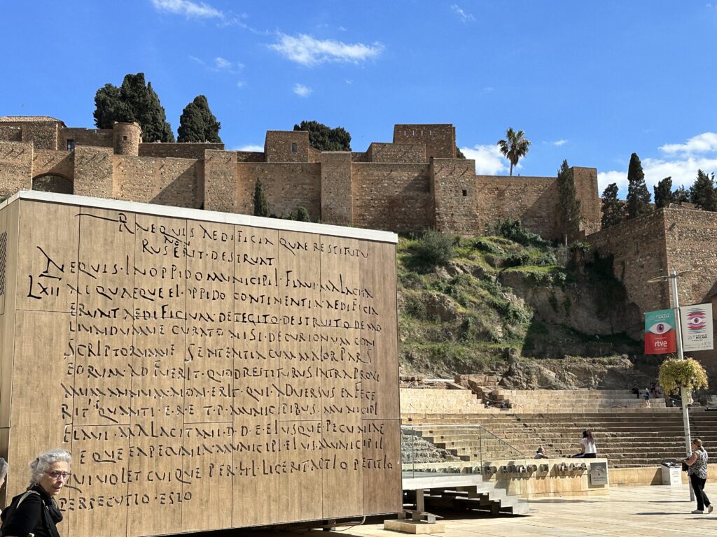 Ancient stone fortress walls rise behind a wooden panel inscribed with Latin text in a Mediterranean plaza