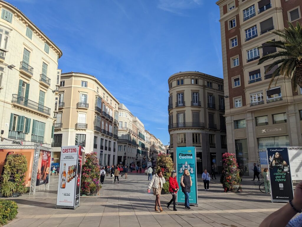 Bustling pedestrian plaza in Malaga with historic architecture, storefronts, and promotional banners under clear blue sky