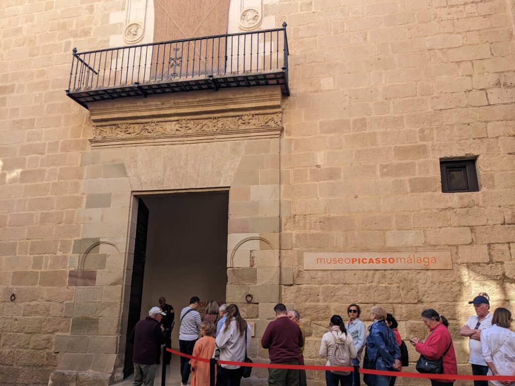 Visitors queue at the entrance of Museo Picasso Málaga, a stone building with ornate architectural details and a wrought-iron balcony
