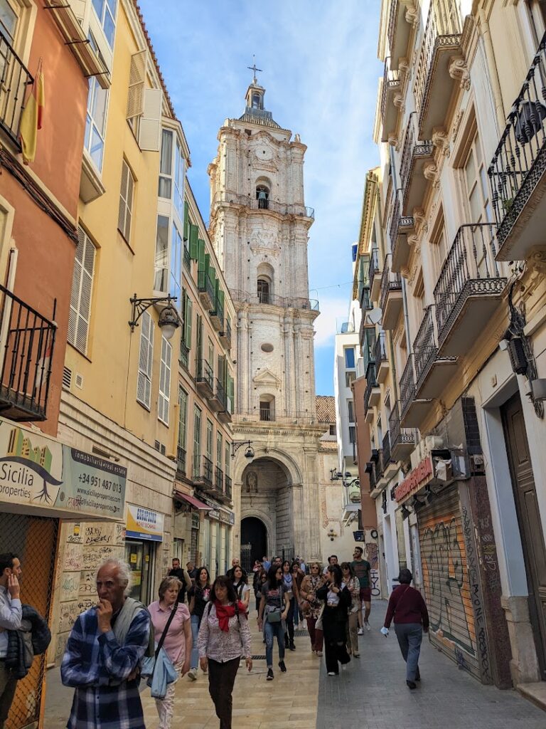 A historic cathedral tower rises above a narrow European street lined with colorful buildings and pedestrians