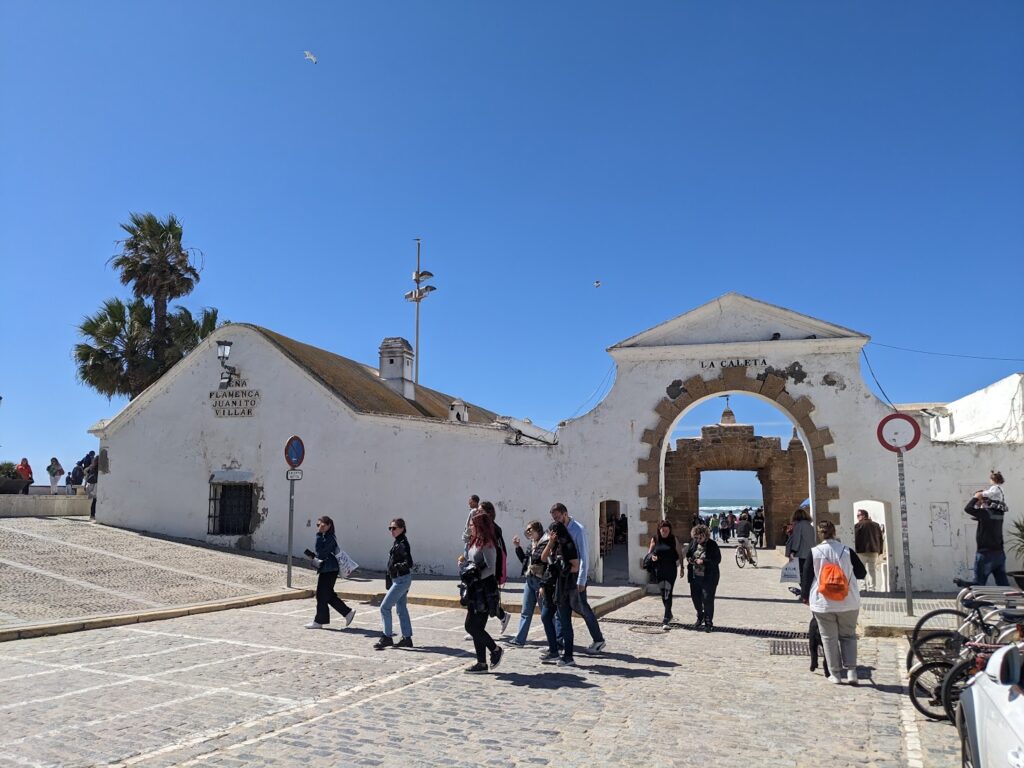 White-walled historic buildings with a large archway entrance at a coastal village, visitors walking in the plaza under clear blue sky