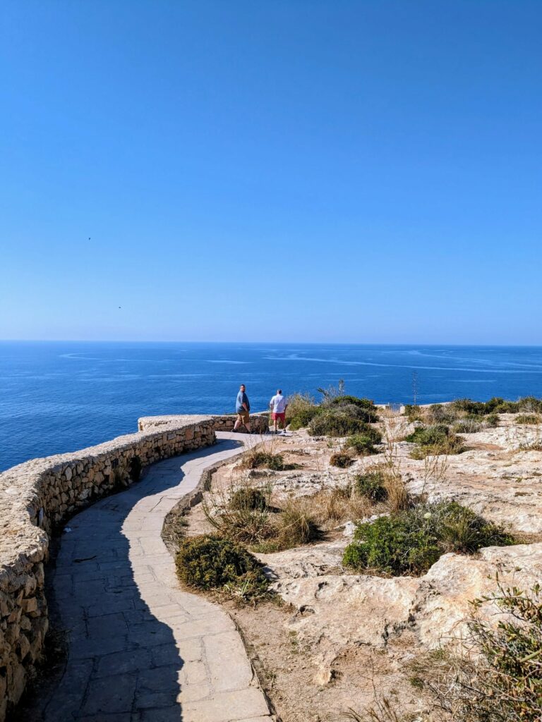 Curving stone path along a rocky coastline with two people overlooking the ocean under a clear sky.