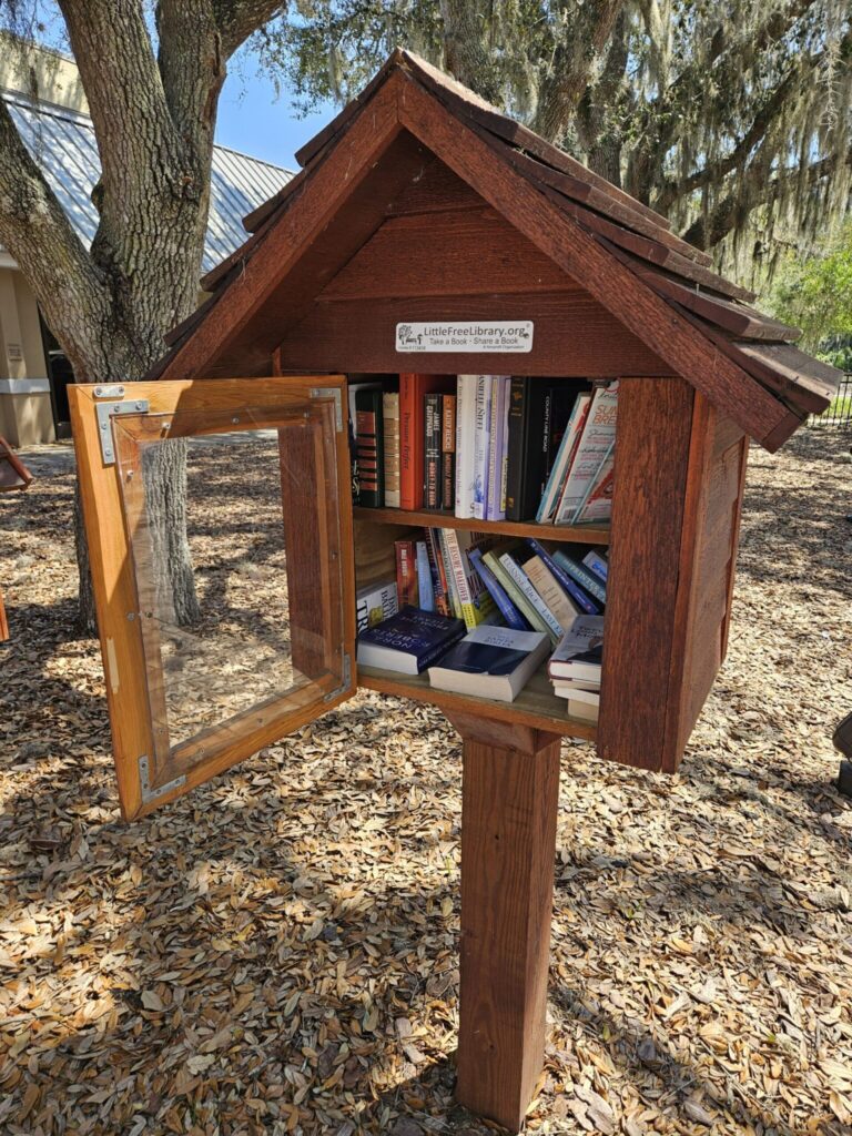 A wooden Little Free Library filled with books stands beneath Spanish moss-draped trees in a sunny outdoor setting