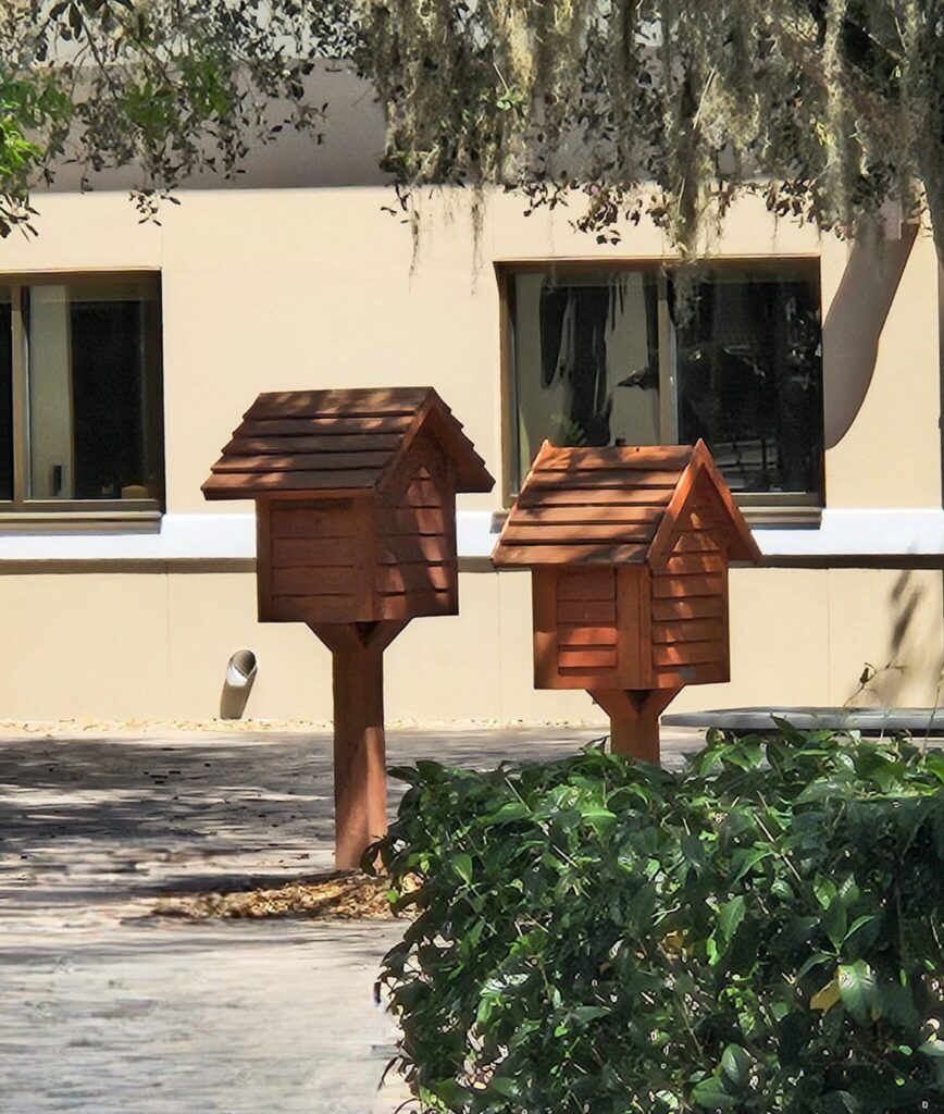 Two wooden birdhouses stand in a sunny courtyard beside a modern white building with large windows