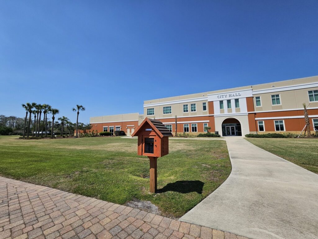 A red mailbox stands on a lawn in front of a large orange and cream-colored City Hall building with palm trees
