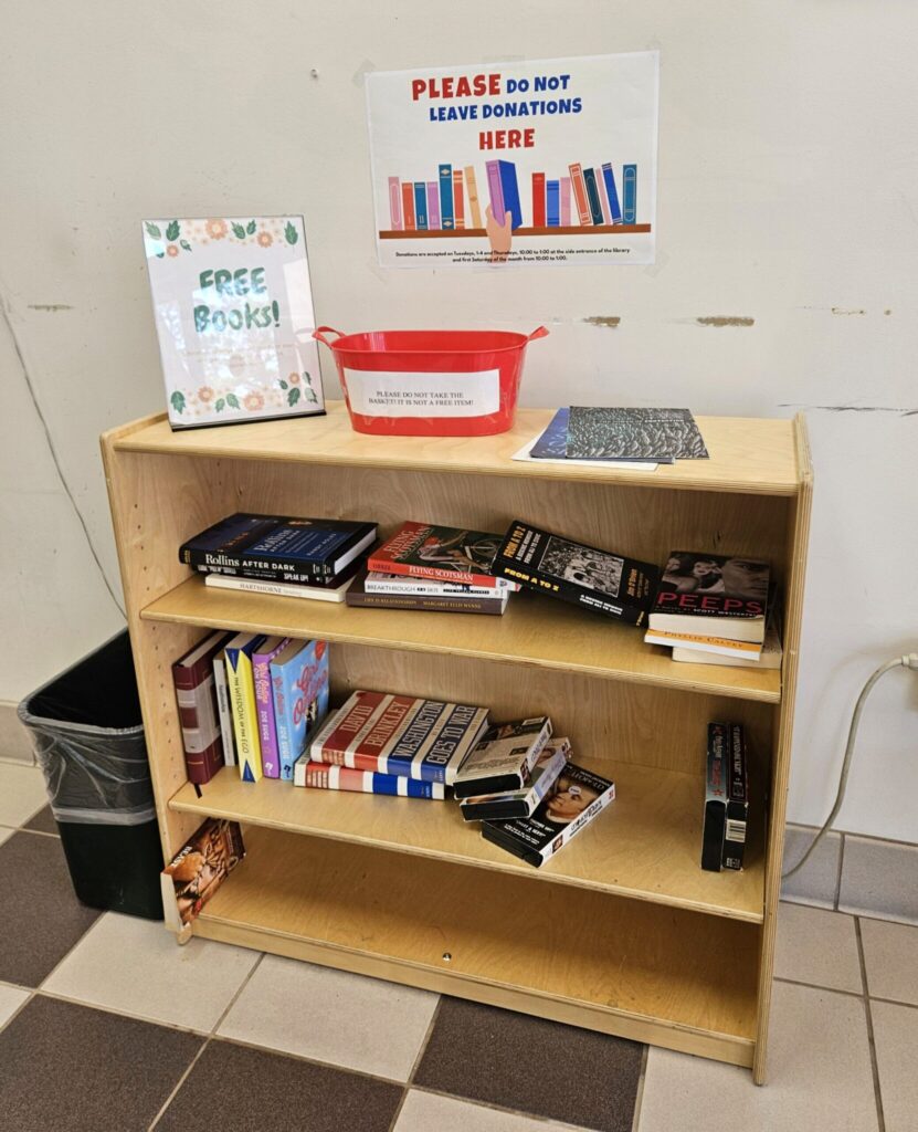 Wooden bookshelf with free books sign and red donation bin against white wall with library signage