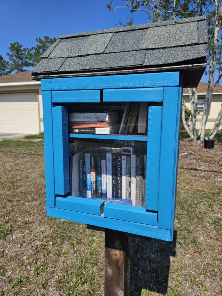 Blue wooden little free library filled with books stands in a residential yard with houses and trees visible in the background
