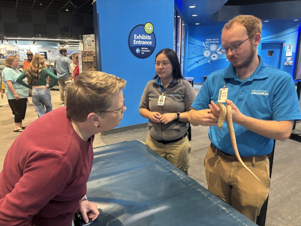Museum staff member demonstrates a snake to a visitor in an exhibits area with blue walls and other guests in background