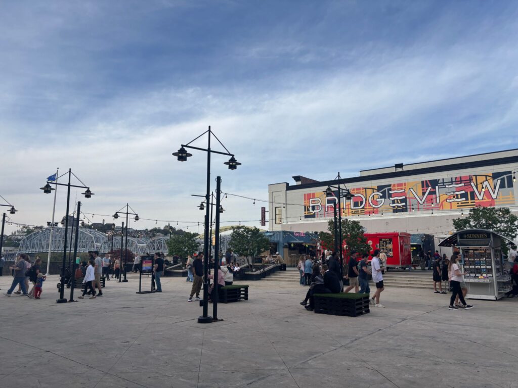 Busy amusement park plaza with vintage-style street lamps, colorful arcade building, and visitors walking on a sunny day