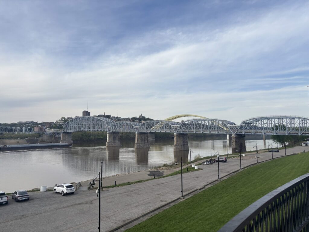 A steel arch bridge spans a wide river with a waterfront promenade and green lawn in the foreground under a partly cloudy sky
