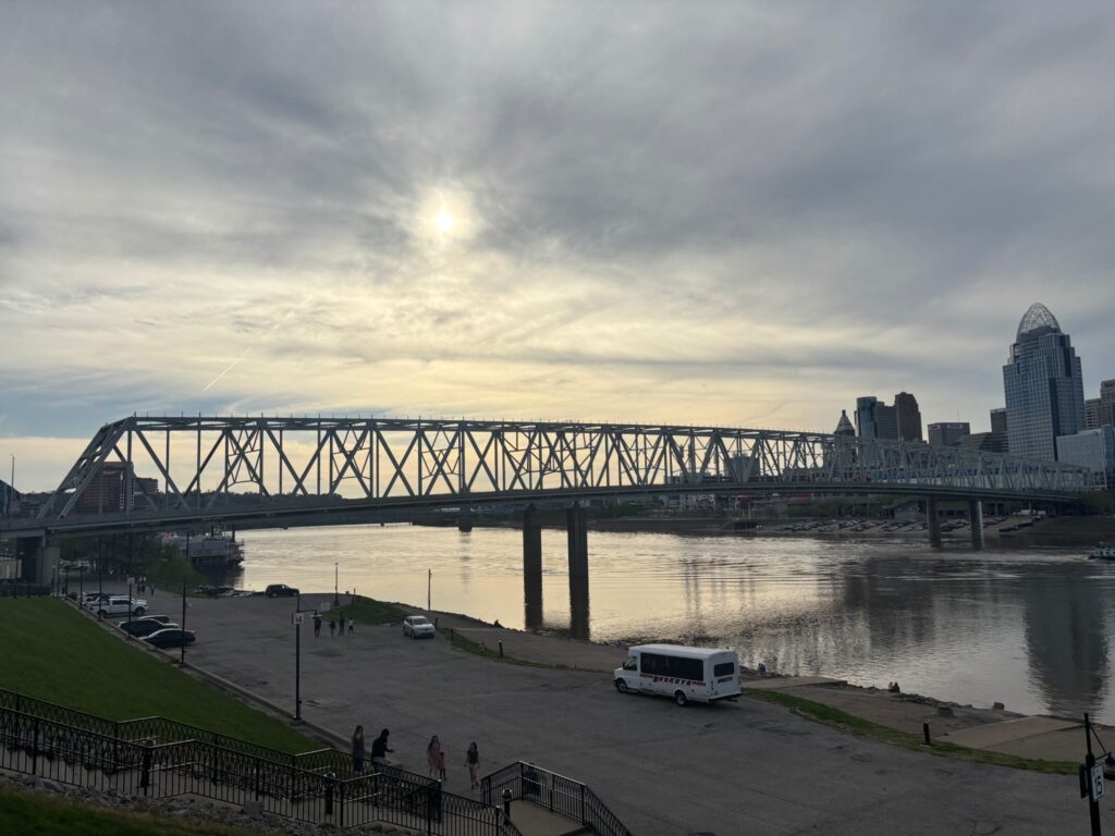 Steel truss bridge spans a river at dusk with Cincinnati skyline and cloudy sky in the background