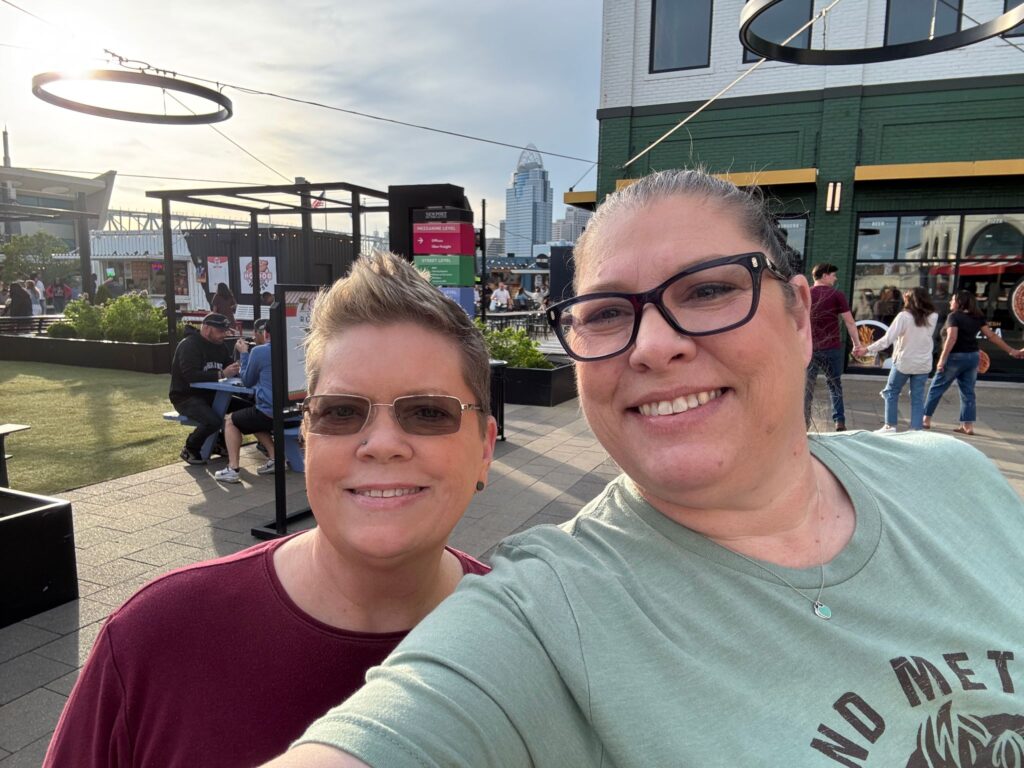 Two women wearing sunglasses smile at the camera in an outdoor urban plaza with buildings and crowds in the background
