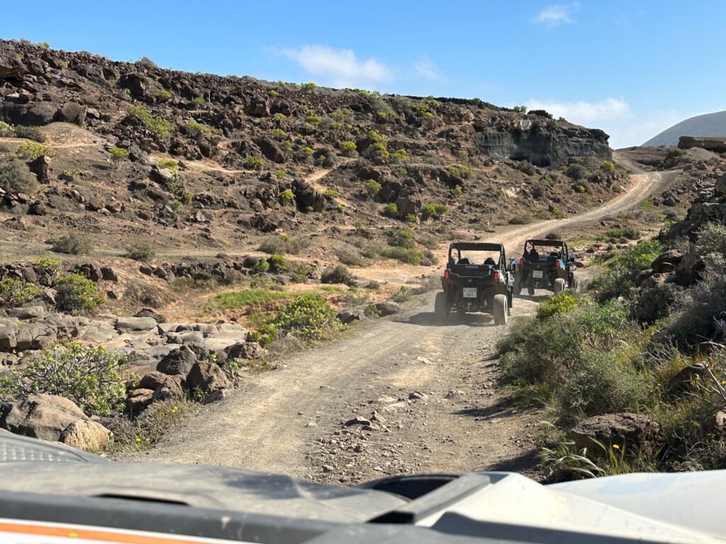Two off-road vehicles navigate a rocky dirt trail through a volcanic landscape with sparse vegetation and rocky hillsides
