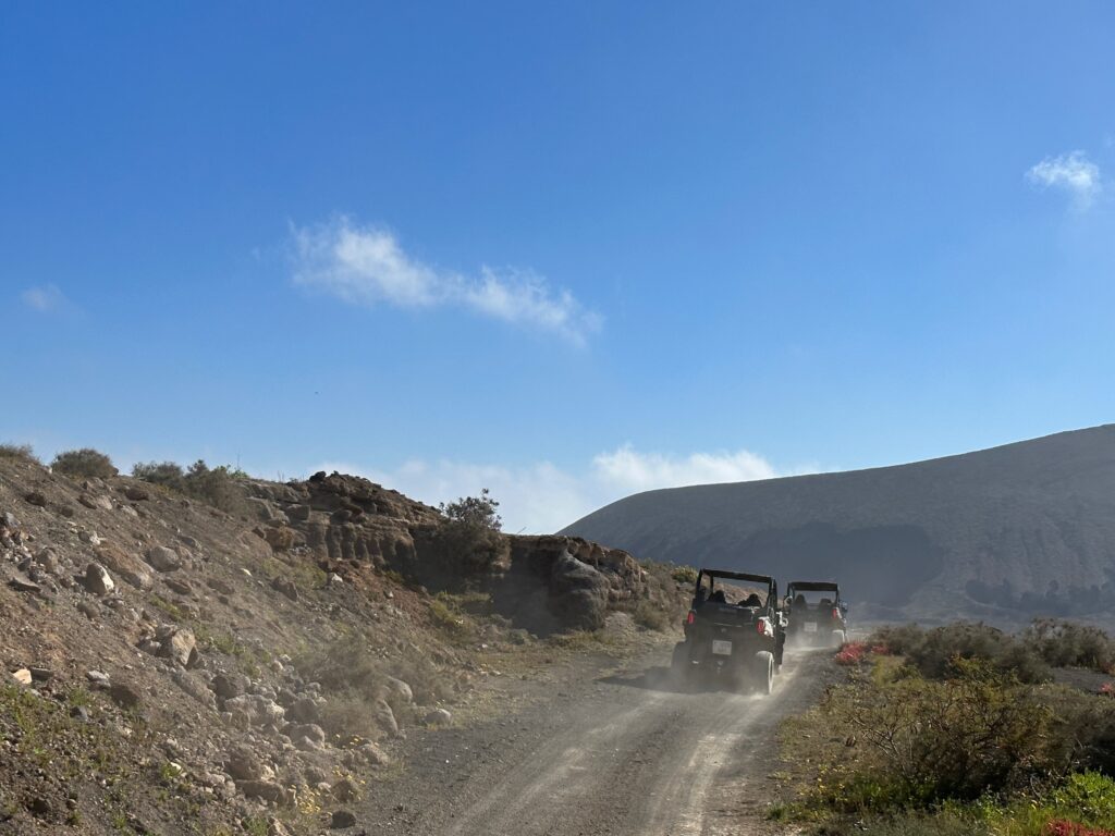 Two rugged vehicles travel on a dusty mountain road through arid terrain under a clear blue sky