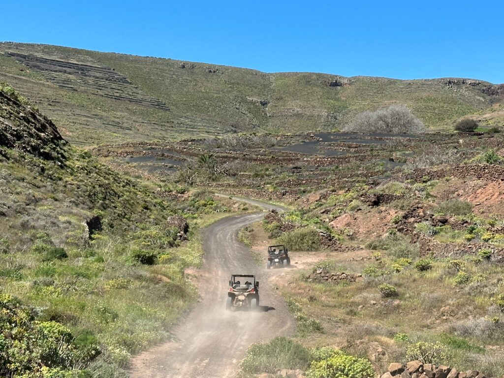 Two off-road vehicles travel down a dusty mountain trail through a semi-arid landscape with sparse vegetation